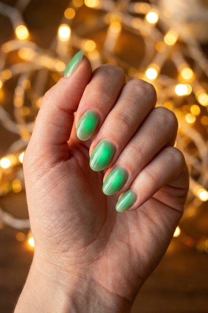 Short almond nails in a soft emerald green aura that glows from the center outward, on a fair-skinned hand near blurred fairy lights.