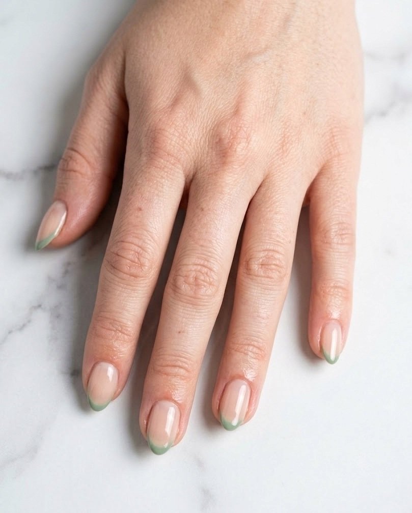 Short almond nails with sheer nude base and thin sage green French tips on a fair skin tone hand