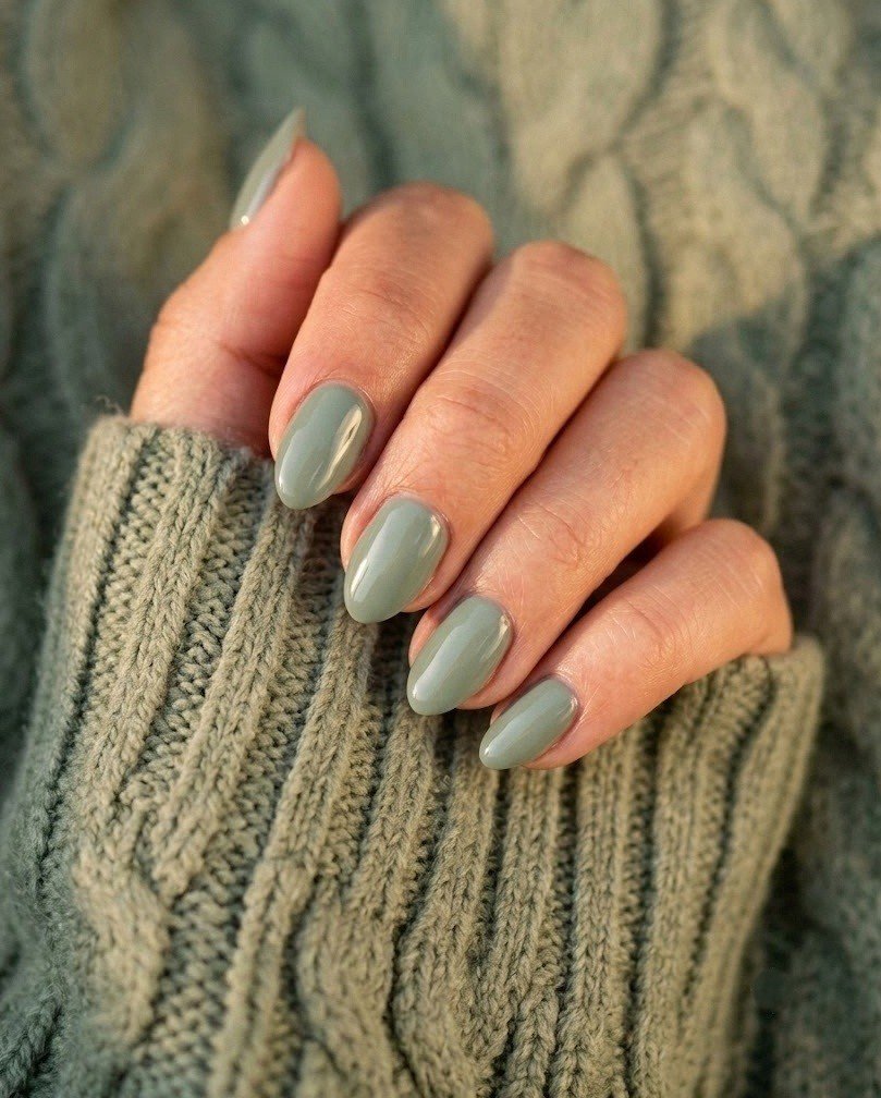 Short soft-almond nails in glossy dusty sage green on a medium skin tone hand against a sage knit sweater