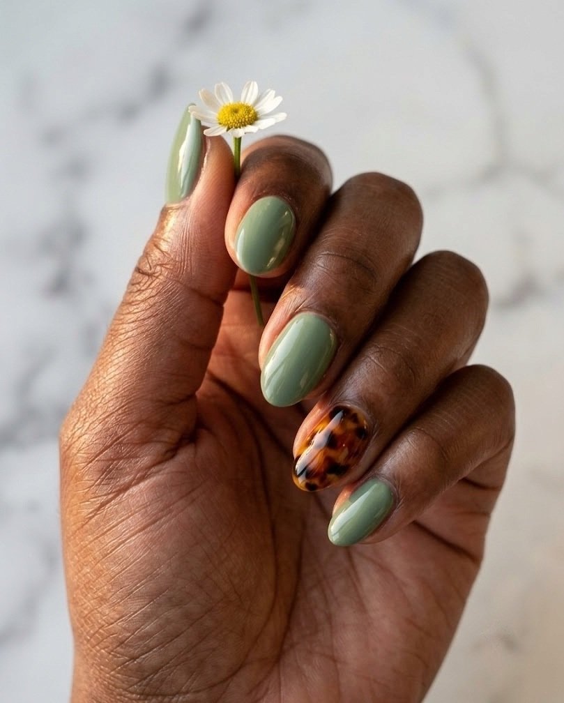 Short almond sage green nails with one amber and brown tortoiseshell accent nail, deep skin tone hand