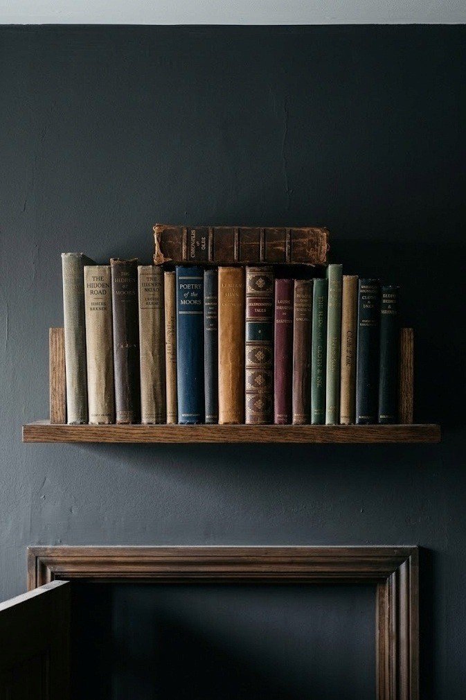 Narrow shelf mounted above a bedroom door frame holding a row of cloth bound and leather bound books against dark walls