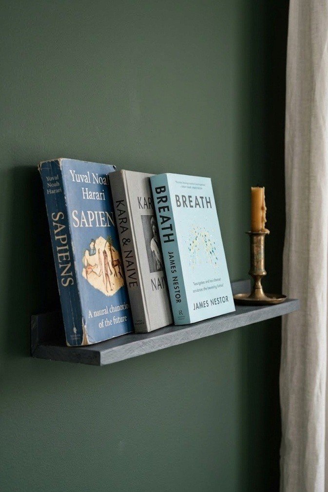 Narrow slate adhesive picture ledge on a deep green wall holding four hardcover books face out and a small brass candlestick