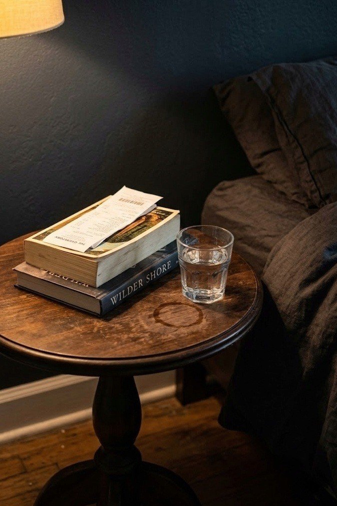 Small round dark wood pedestal side table beside a bed holding two stacked books and a short glass of water