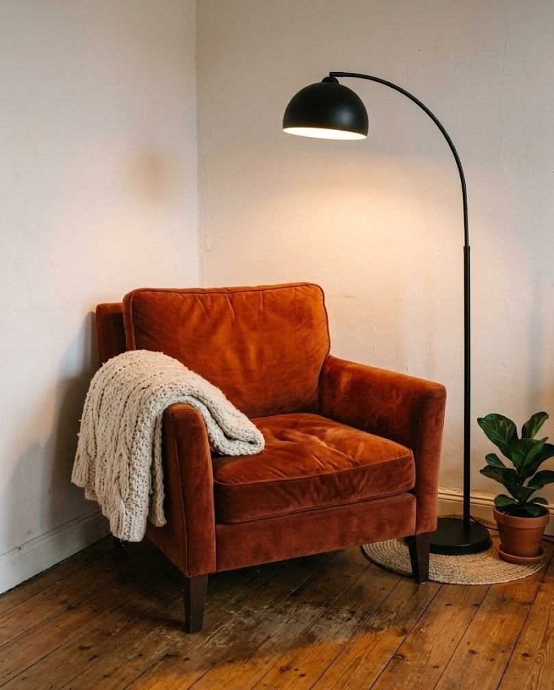 Rust burnt-orange velvet armchair in a small apartment corner with a cream knit throw and a black arc lamp